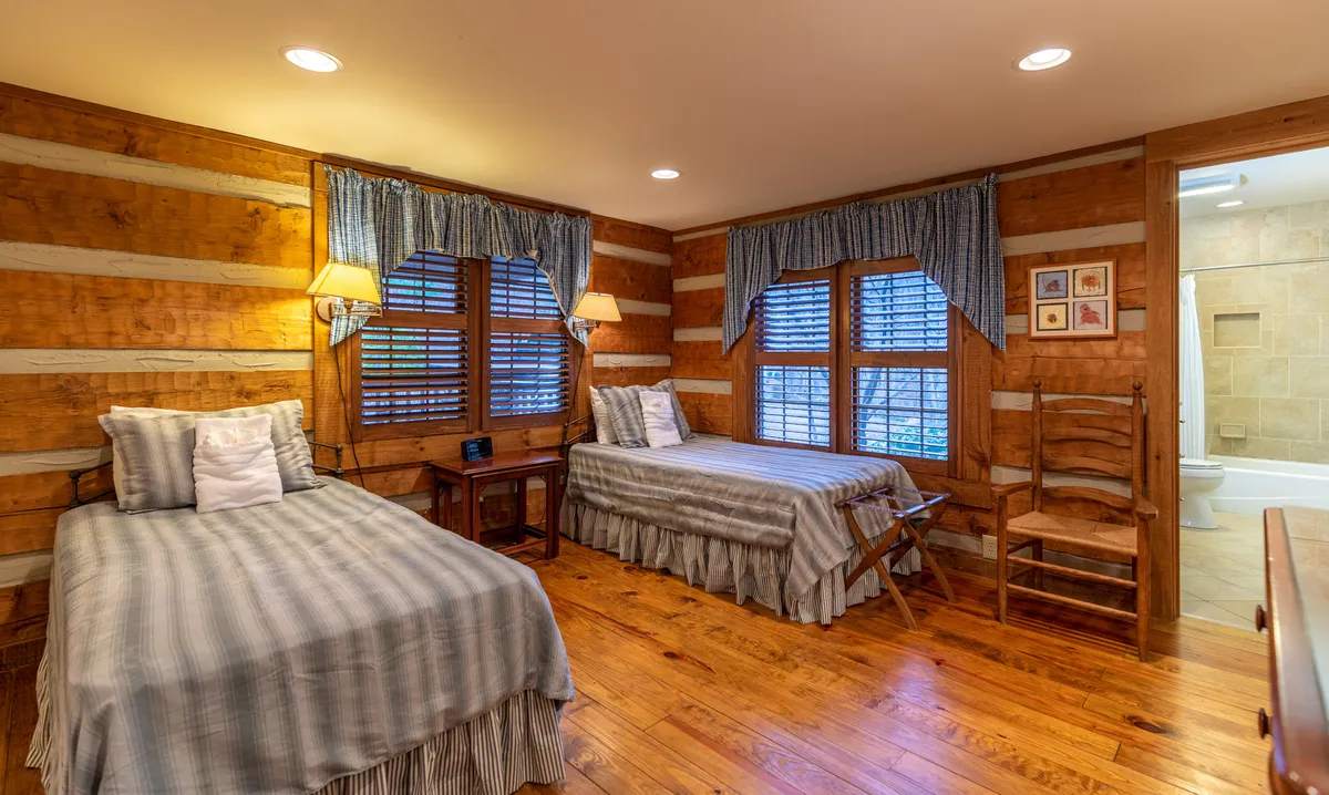 A bedroom with two twin beds, wooden furniture, and large windows with shutters that let in abundant morning light. The scene exudes a welcoming, homely feel ideal for families or groups.