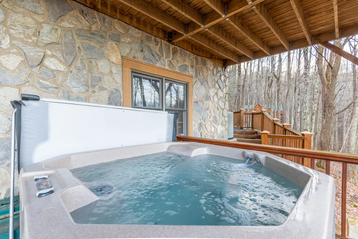 This image shows a tranquil wooden deck outfitted with a hot tub, bordered by a rustic stone wall. The deck is surrounded by natural greenery, conveying a strong sense of immersion in the outdoors. The space looks inviting for both people and pets, with ample room to lounge and take in the fresh mountain air.