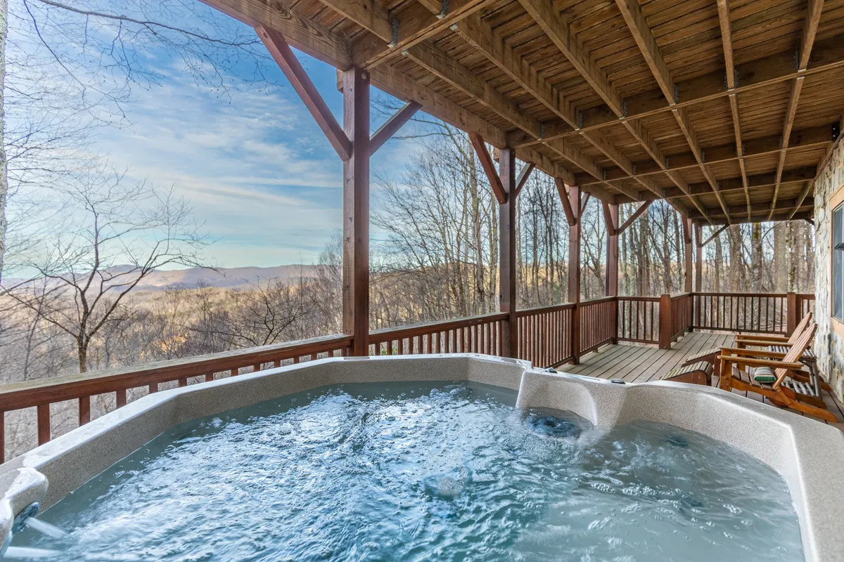 An outdoor hot tub sits on a spacious wooden deck, surrounded by natural greenery and mountain views. The area invites guests to relax in the hot tub while soaking up the scenery or socializing on the deck.
