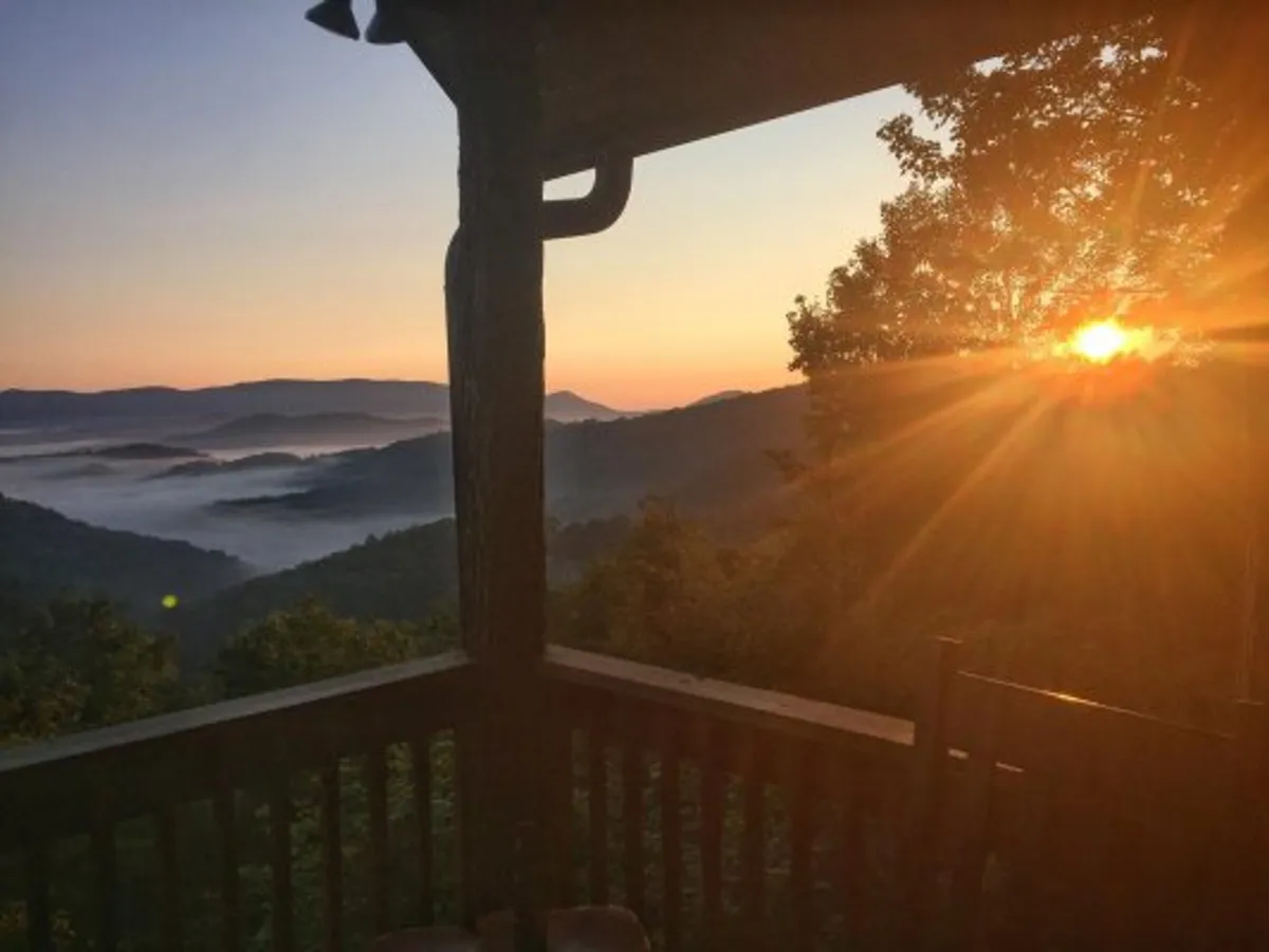 A sunrise scene over mountains framed by a wooden porch railing. The image suggests a peaceful start to the day, potentially enjoyed with coffee or quiet conversation, as the landscape glows with early morning light.