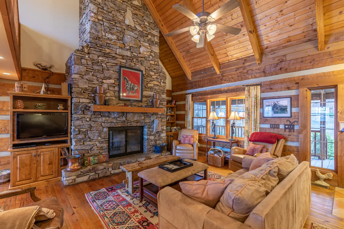 A spacious Great Room featuring a massive stone fireplace, exposed wooden beams, cozy seating arrangements, and expansive windows that flood the space with natural light and frame sweeping mountain vistas. The scene is set for family gatherings, relaxation by the fire, or enjoying a sunrise through the windows.