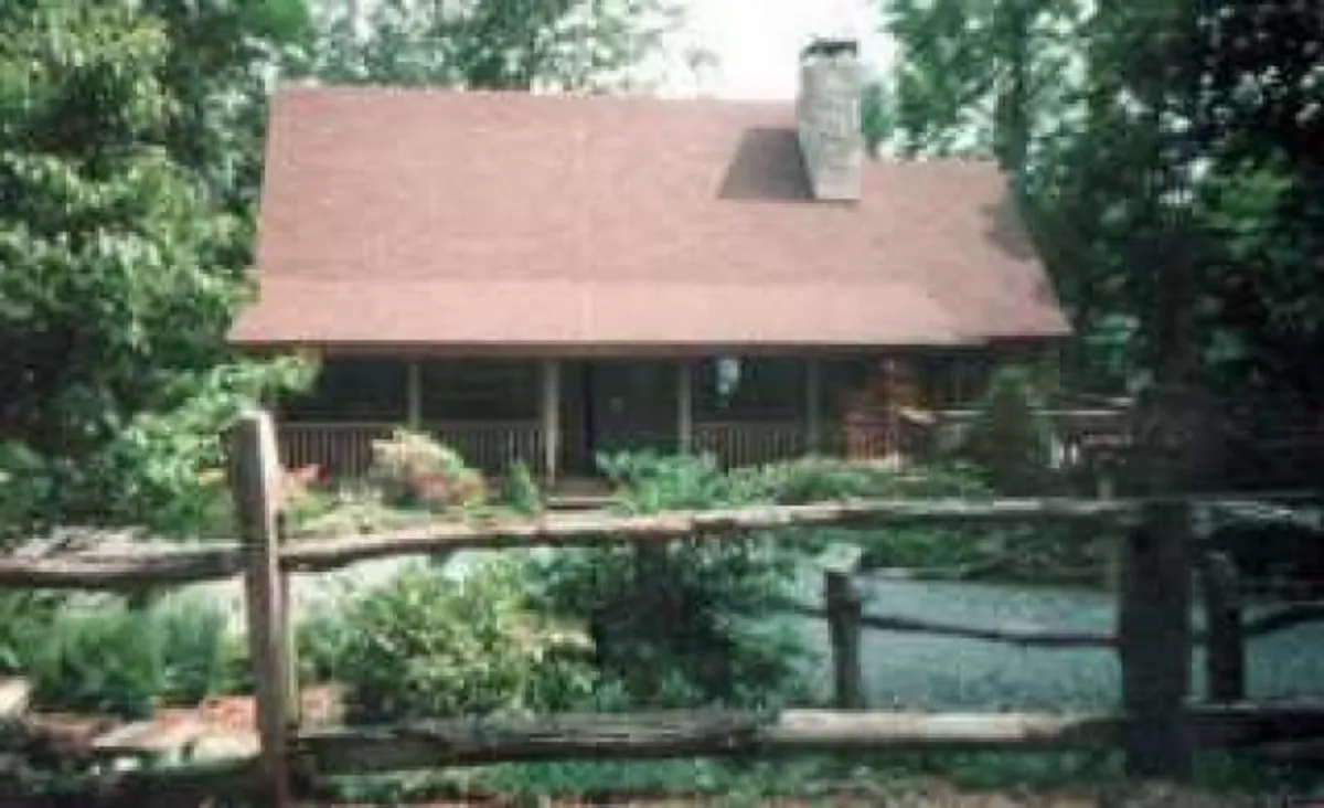 A welcoming view of the property's exterior, featuring a wooden fence, charming front porch, large chimney, and landscaped garden. The cabin is set amidst lush greenery, suggesting plenty of open space for dogs to explore and for guests to enjoy the mountain atmosphere from the porch.