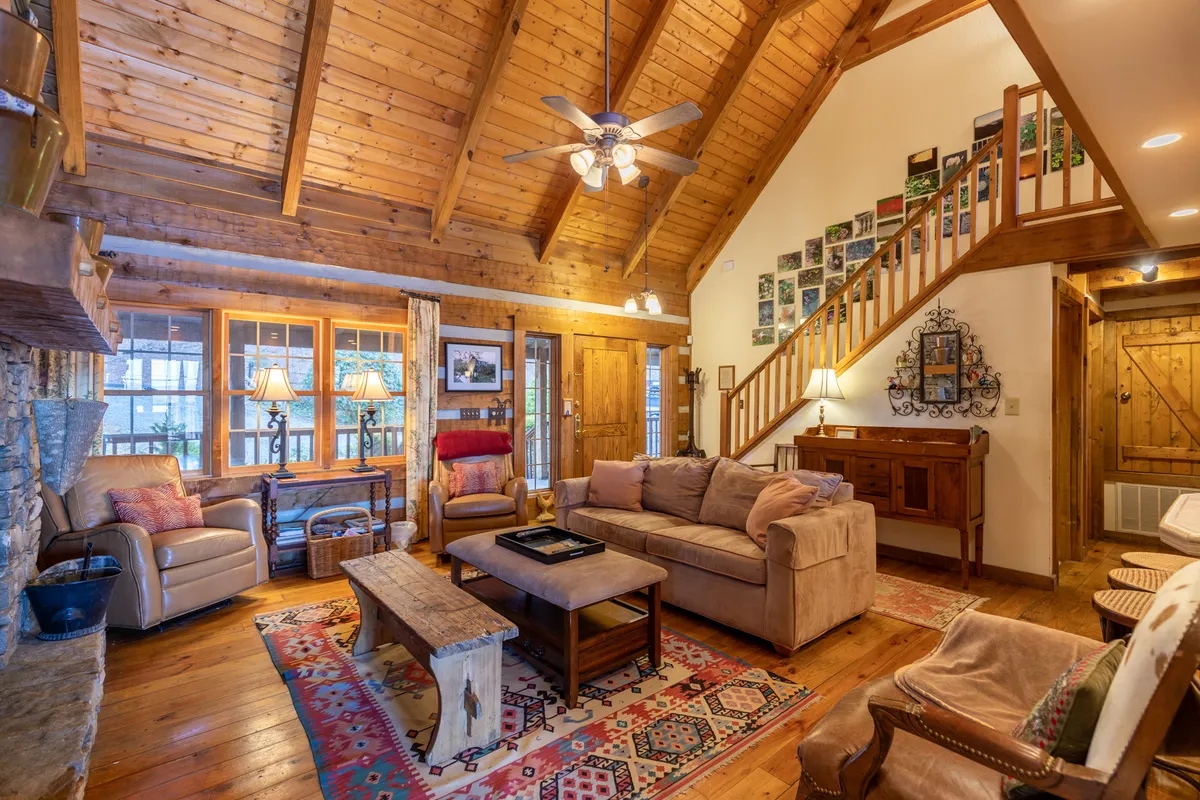 A welcoming living space featuring rugged wooden ceiling beams and a prominent stone fireplace. Comfortable seating is arranged around the fire, with a decorative rug anchoring the scene, creating a cozy atmosphere perfect for gathering during colder months.