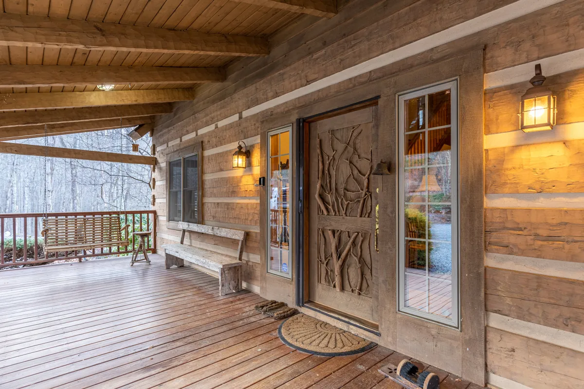 A cozy and rustic front porch featuring an intricately carved wooden door, a classic swing bench, and warm wall sconces. Large windows frame the inviting entrance, hinting at the home’s welcoming atmosphere and the promise of memorable moments awaiting inside.