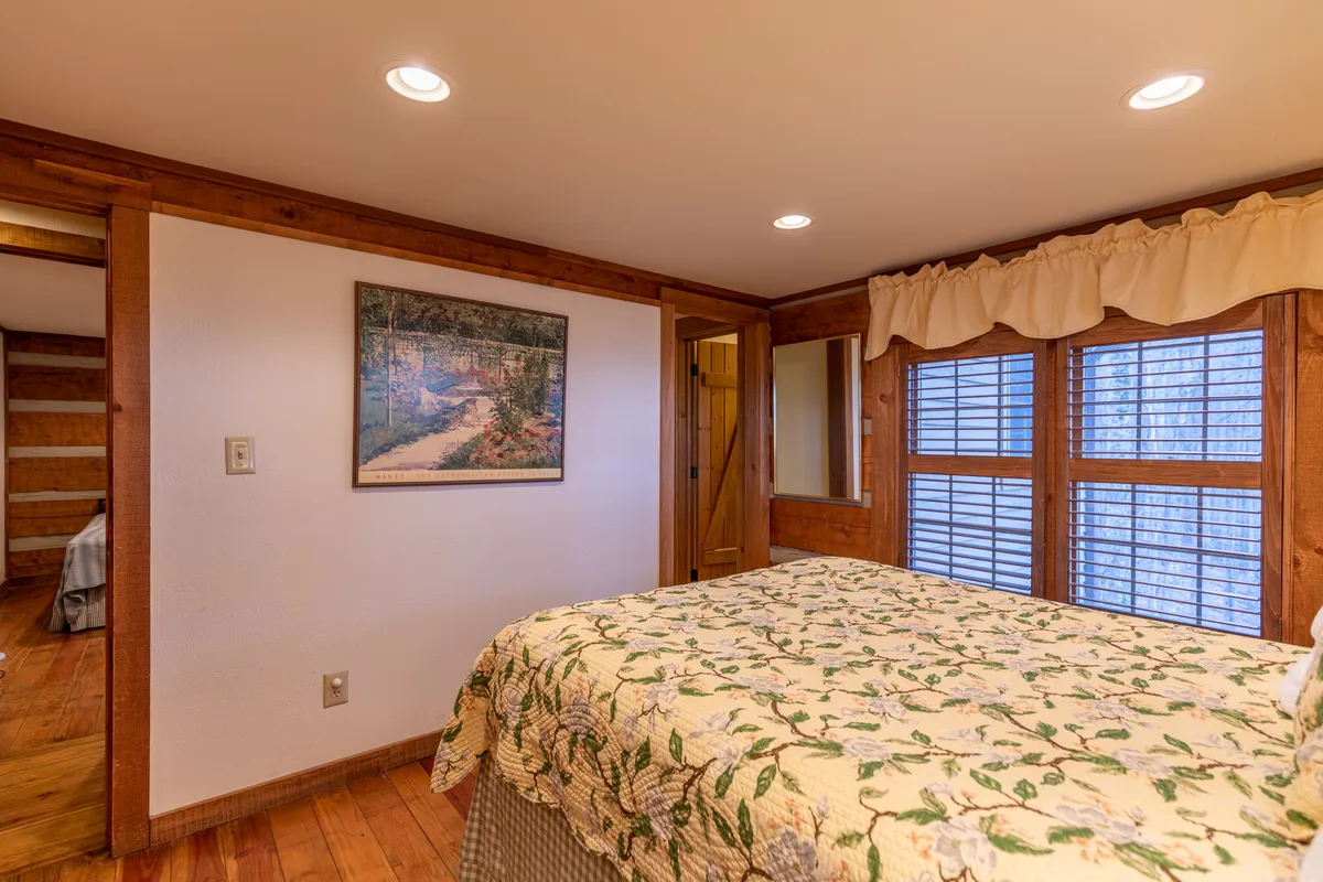 A warmly lit bedroom with a quilted bedspread, wooden window frames, a framed painting, and a mirror. The natural light streaming through the windows adds to the inviting atmosphere of the space.