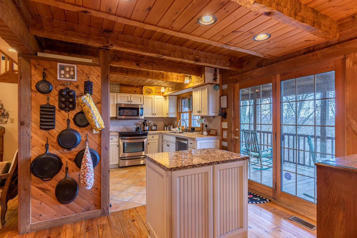 This image showcases a modern, fully equipped kitchen with a granite countertop, wooden beams, and hanging pots and pans. Double glass doors bring in natural light, and the space exudes a cozy mountain charm ideal for both cooking and entertaining.