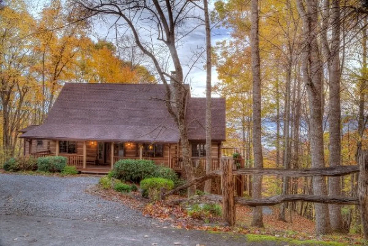 The image showcases a charming log cabin nestled among vibrant autumn foliage, with a rustic wooden fence in the foreground. The scene feels warm and inviting, suggesting opportunities to relax on the porch, take in sweeping mountain views, and enjoy crisp mountain air. The overall mood is cozy and tranquil, perfectly capturing the allure of a private mountain getaway in the fall.