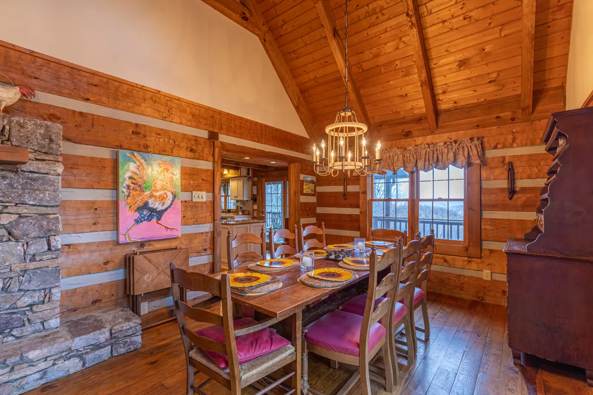 A warm, inviting dining room with a large table set for gatherings, a glowing stone fireplace that connects visually to the Great Room, elegant chandelier lighting, and windows that offer scenic views. Artwork and decor add to the space's welcoming ambiance, perfect for memorable family meals.