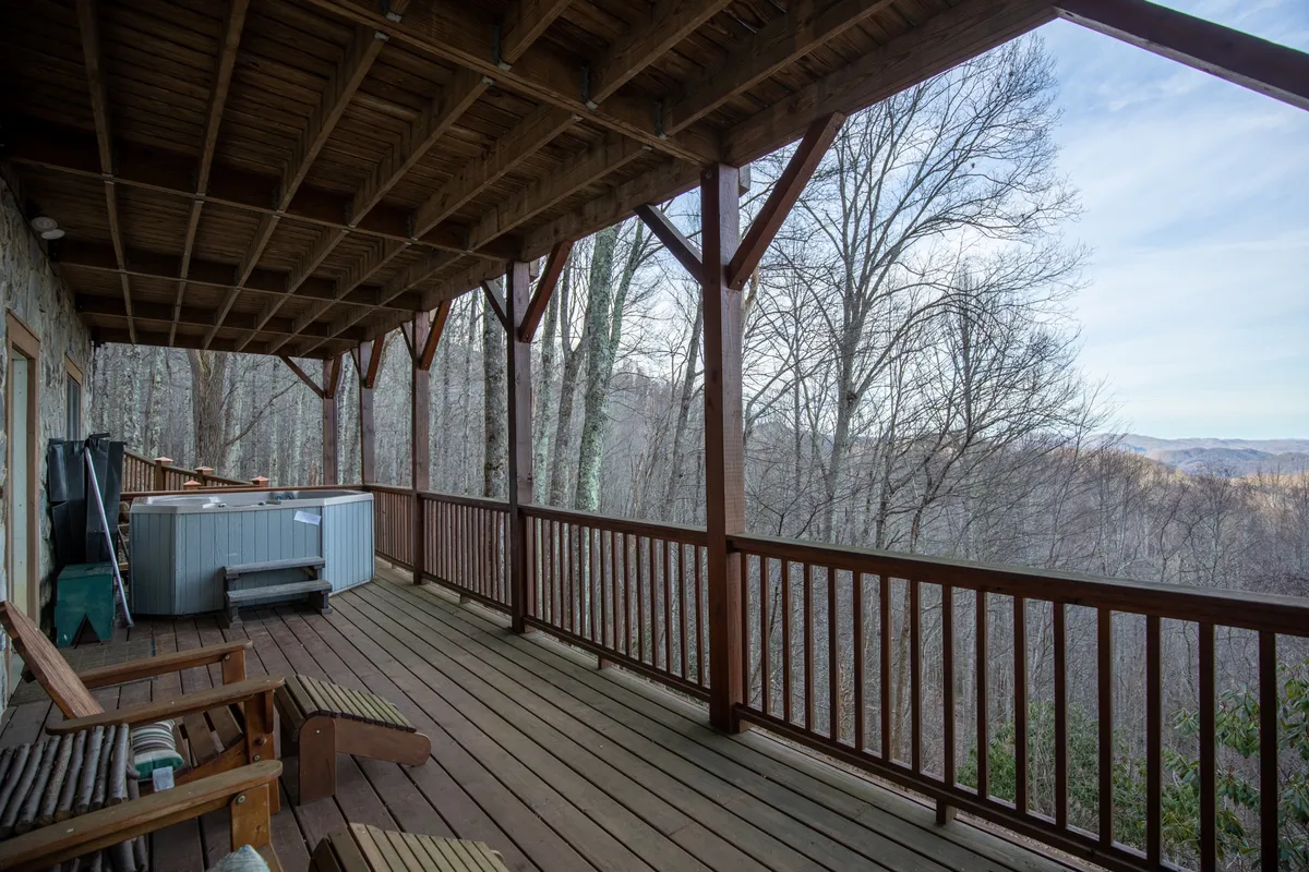 An inviting exterior scene featuring a wooden wrap-around deck equipped with a private hot tub and comfortable deck chairs, all overlooking a sweeping panorama of distant mountains. The scene is serene, suggesting family relaxation and group gatherings with nature as the backdrop.